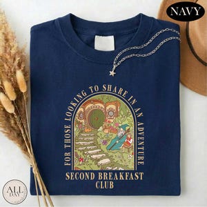 A navy blue t-shirt with a graphic design depicting a castle or palace surrounded by greenery, with the text "For those looking to share in an adventure" and "Second Breakfast Club" printed on it. The t-shirt is folded and placed on a white surface, with some dried flowers and a brown hat visible in the background.