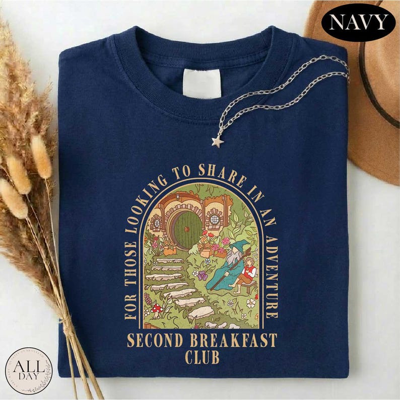 A navy blue t-shirt with a graphic design depicting a castle or palace surrounded by greenery, with the text "For those looking to share in an adventure" and "Second Breakfast Club" printed on it. The t-shirt is folded and placed on a white surface, with some dried flowers and a brown hat visible in the background.