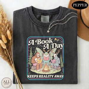 May include: A charcoal gray t-shirt featuring a graphic print of a Bigfoot, a moth creature, and a deer-headed figure sitting around a campfire reading books. The text reads A BOOK A DAY KEEPS REALITY AWAY. The design includes trees and stars.
