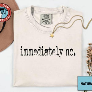 May include: Cream-colored t-shirt with the phrase "immediately no." printed in black. The shirt has a crew neck and a tag that reads "Comfort Colors." A gold necklace with a star pendant rests on the shirt.