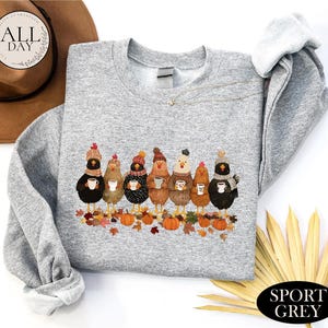 May include: A sport grey crewneck sweatshirt featuring a graphic of seven chickens wearing winter hats and scarves, each holding a mug. The chickens stand among fallen autumn leaves and orange pumpkins. The sweatshirt has long sleeves and a ribbed collar.