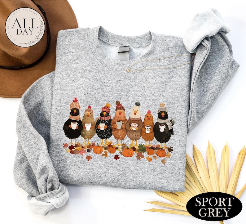 May include: A sport grey crewneck sweatshirt featuring a graphic of seven chickens wearing winter hats and scarves, each holding a mug. The chickens stand among fallen autumn leaves and orange pumpkins. The sweatshirt has long sleeves and a ribbed collar.