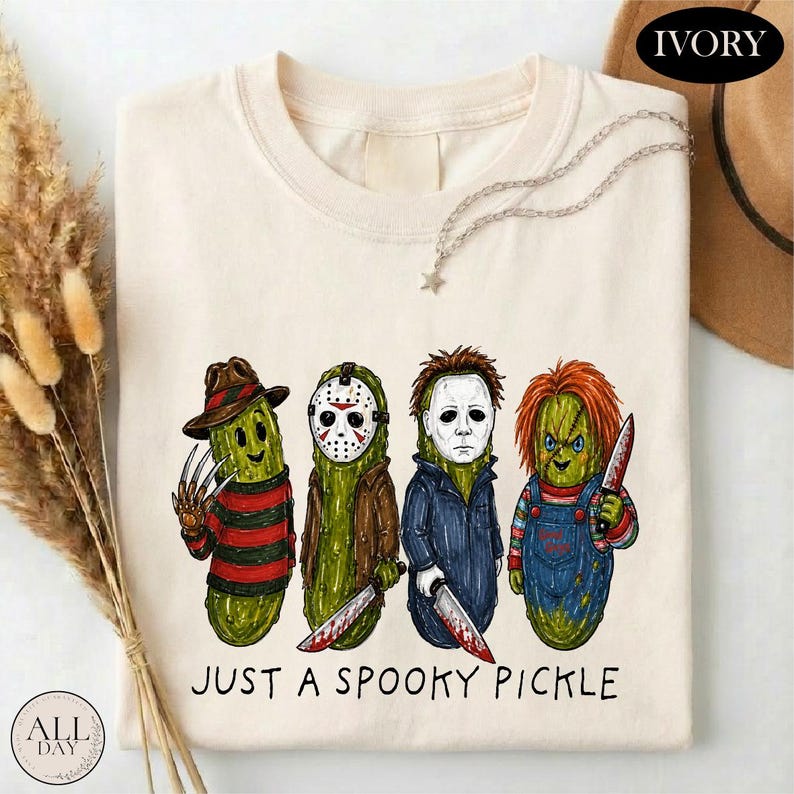 May include: An ivory t-shirt featuring a graphic of four green pickles dressed as horror movie characters, each holding a weapon. Below the characters, the text reads JUST A SPOOKY PICKLE in black lettering. The shirt has a crew neckline.