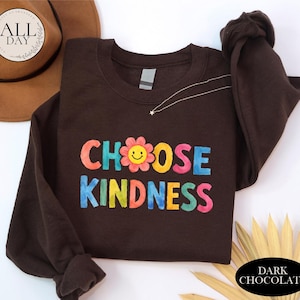 A brown sweatshirt with the text "CHOOSE KINDNESS" printed on it, along with a flower design. The sweatshirt is placed on a white surface, and there is a brown hat and some leaves in the background.