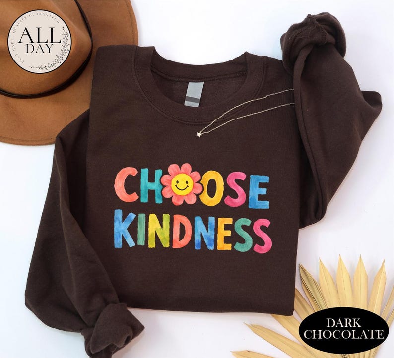 A brown sweatshirt with the text "CHOOSE KINDNESS" printed on it, along with a flower design. The sweatshirt is placed on a white surface, and there is a brown hat and some leaves in the background.