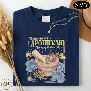 a navy blue t-shirt with a graphic design depicting a mortar and pestle surrounded by flowers and herbs, along with the text "Mama's Apothecary" and "Rejuvenates Medicine Poison". The t-shirt is displayed on a white surface, with a brown hat and dried flowers in the background.