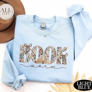 A light blue sweatshirt with the word "BOOK" printed on it, along with the word "Worm" written below it. The sweatshirt is placed on a white background, and there is a brown hat and some straw in the background.