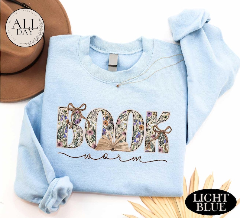 A light blue sweatshirt with the word "BOOK" printed on it, along with the word "Worm" written below it. The sweatshirt is placed on a white background, and there is a brown hat and some straw in the background.