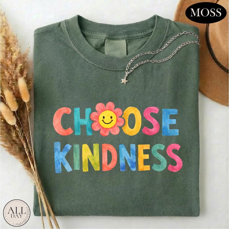 A green t-shirt with the text "CHOOSE KINDNESS" printed on it, along with a flower design. The t-shirt is folded and placed on a white surface, with some dried flowers and a hat nearby.