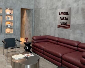Può includere: Interno di un moderno soggiorno con un divano componibile in pelle bordeaux, un tavolino in acciaio inossidabile e una sedia nera. Un'opera d'arte incorniciata sulla parete recita "AMORE PASTA VINO". La stanza ha un design minimalista con pareti in cemento.