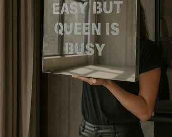 May include: A square mirror with the text "LOVE IS EASY BUT QUEEN IS BUSY" in silver glitter lettering. The mirror is held up by a person wearing a black shirt and black pants.