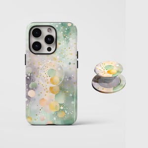 May include: A smartphone case and a matching circular collapsible phone grip featuring an abstract watercolor pattern in shades of sage green, soft peach, and gold speckles. The case has a cutout for a triple-lens camera system.