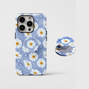 Pode incluir: Uma capa de telefone azul claro e um suporte pop-socket a condizer, ambos com um padrão de margaridas com pétalas brancas e centros amarelos. A capa foi concebida para um smartphone, e o suporte pode ser usado como pega ou suporte.