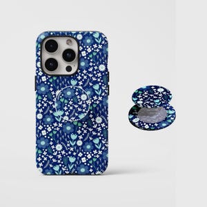 Pode incluir: Uma capa de smartphone azul-escura e um suporte circular retrátil para telemóvel a condizer, ambos com um padrão repetitivo de pequenas flores, folhas e caules brancos, azul-claros e verde-azulados em toda a superfície dos acessórios.