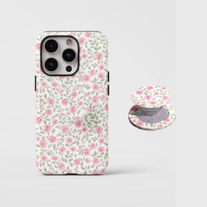 Pode incluir: Uma capa de telefone branca e um suporte de telefone com um padrão de rosas cor-de-rosa e folhas verdes. A capa tem um módulo de câmera preto e botões laterais pretos. O suporte é redondo e dobrável.