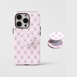 May include: A pink phone case and matching phone grip featuring a repeating pattern of white skulls and hearts. The phone case is on a white surface. The phone grip is detached and next to the phone case.
