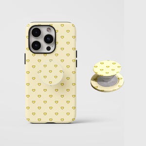 Pode incluir: Uma capa de smartphone amarelo-claro com um padrão repetitivo de pequenos corações amarelos com contornos escuros, acompanhada por um suporte circular dobrável para telemóvel a condizer que exibe o mesmo padrão de corações na superfície.