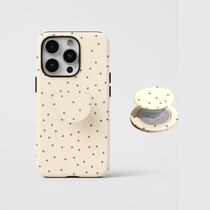 Pode incluir: Uma capa de smartphone na cor creme e um suporte circular retrátil para telemóvel a condizer, ambos com um padrão de pequenos pontos roxos escuros uniformemente espaçados. A capa tem um recorte para um sistema de câmara tripla.