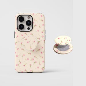 Pode incluir: Uma capa de smartphone na cor creme com um padrão repetitivo de pequenas flores cor-de-rosa com caules verdes, acompanhada por um suporte circular dobrável para telemóvel a condizer que exibe o mesmo design floral na superfície plana.