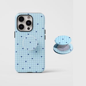 May include: A light blue phone case and matching phone grip featuring a grid pattern with small navy blue hearts. The phone case is designed to fit a smartphone, and the grip is attached to the back.