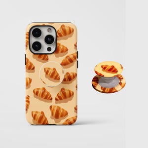Puede incluir: Una funda y un soporte para teléfono con un patrón repetido de cruasanes dorados sobre un fondo beige claro. La funda del teléfono tiene un módulo de cámara negro y botones laterales negros. El soporte del teléfono es redondo y tiene un diseño de cruasán a juego.