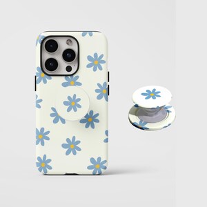 Pode incluir: Uma capa de telefone e um suporte a condizer com um padrão de margaridas azuis e amarelas sobre um fundo creme. A capa é projetada para um smartphone, e o suporte é fixado na parte de trás.