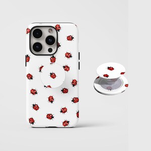 May include: White phone case and pop socket with a repeating pattern of red ladybirds with black spots. The phone case has a camera cutout and black side accents. The pop socket is round and collapsible.