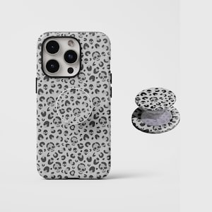 Pode incluir: Uma capa de smartphone cinza-claro e um suporte circular retrátil para telefone a condizer, ambos com um padrão repetitivo de manchas de leopardo cinza-escuro intercaladas com pequenos ícones de caveiras em toda a superfície.