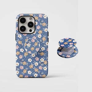 Pode incluir: Uma capa de smartphone azul e um suporte circular para telemóvel a condizer com um padrão repetitivo de pequenas flores brancas, cor-de-rosa e bege com folhas verdes e pontos brancos sobre um fundo azul. A capa tem um recorte para a lente.