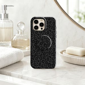 Può includere: Una custodia per telefono con stampa leopardata nera e grigio scuro, dotata di un'impugnatura circolare pieghevole sul retro. La custodia presenta un'apertura per l'obiettivo della fotocamera e copre il retro e i lati del dispositivo.