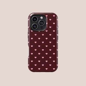 May include: A maroon phone case with a repeating pattern of small pink hearts. The case has a glossy finish and a camera module with three lenses. The phone case is designed to protect the phone from scratches and damage.