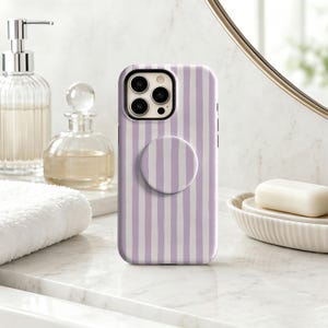 May include: A smartphone case featuring a vertical lavender and white striped pattern with a matching circular collapsible grip attached to the center back, showcasing a triple-lens camera cutout on the upper left corner.