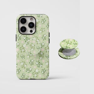 Pode incluir: Uma capa de smartphone verde sálvia e um suporte circular retrátil a condizer. Ambos os artigos apresentam um padrão repetitivo de pequenas flores brancas, folhas verdes e minúsculas flores cor-de-rosa claro em toda a superfície.