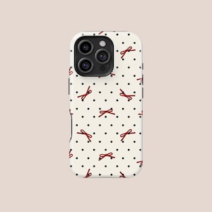 Op de afbeelding: Een witte telefoonhoes met een patroon van zwarte stippen en rode strikken. De hoes heeft een uitsparing voor een drievoudige cameralens en een zwarte rand. Het ontwerp is eenvoudig en elegant.
