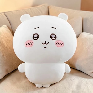 May include: A white plush toy shaped like a round bear with small ears. The bear has embroidered black eyes, a small mouth, and pink blush marks on its cheeks. The toy is sitting on a beige surface.