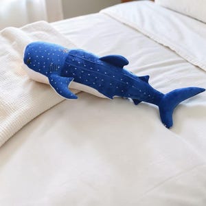 May include: A plush whale shark pillow in shades of blue and white. The soft toy features a dark blue body with white spots, fins, and a tail. It is lying on a white bedspread and a cream-colored blanket.