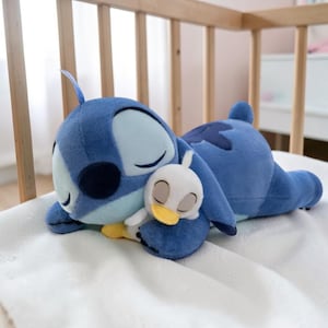 May include: A plush blue Stitch character doll lying on a white blanket, hugging a small white and yellow plush duck. The Stitch doll has large ears and closed eyes, suggesting sleep. The background includes a wooden crib.