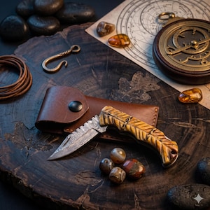 May include: A folding pocket knife with a Damascus steel blade and a carved, feather-textured handle made of brown and tan material. The knife is positioned next to a brown leather sheath with a black snap button closure.
