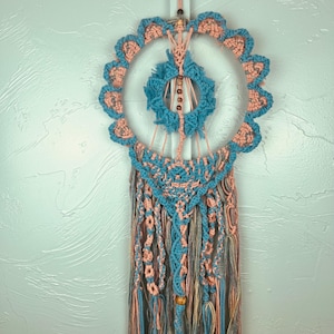 May include: A macrame wall hanging featuring a large circular frame with scalloped peach and blue cord edges. A smaller central ring contains wooden beads on vertical cords. The bottom section has intricate knotted patterns and long hanging tassels.