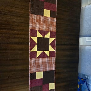 May include: A rectangular quilted table runner featuring a central eight-pointed star block in yellow and dark brown, flanked by sections of brown and white plaid fabric, and bordered by alternating blocks of yellow, dark brown, and maroon fabric.