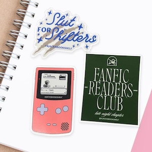 May include: A collection of stickers with book-themed designs. One sticker reads "Slut for Shifters" with a blue and white design. Another is a pink sticker resembling a handheld game console. The third is a green sticker that says "Fanfic Readers Club".