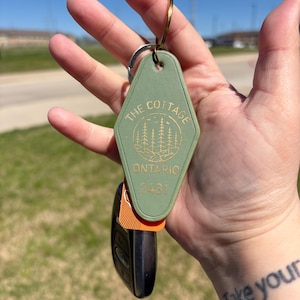 May include: A sage green motel key tag with the words "THE COTTAGE ONTARIO 2481" in gold lettering. The tag is attached to a gold key ring and a black car key. The background is a sunny day with a grassy area.