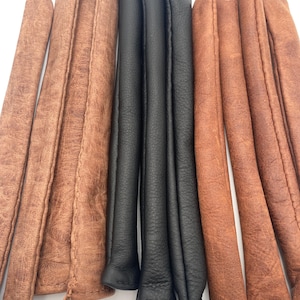 May include: A collection of long, cylindrical, padded covers arranged vertically. The set includes several pieces with a textured, light brown, weathered finish and three pieces with a smooth, matte black finish, all featuring stitched seams.