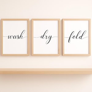 May include: Three framed prints with the words "wash," "dry," and "fold" in black script. The prints have a white background and are set in light brown wooden frames. A wooden shelf is below the prints.