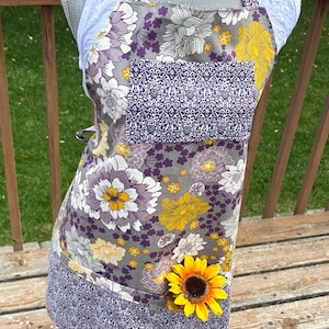 May include: A floral apron with a gray background, featuring large white, yellow, and purple flowers. A purple pocket with a decorative pattern is centered on the chest. A yellow sunflower is attached to the apron's lower right side. The apron is displayed on a mannequin.