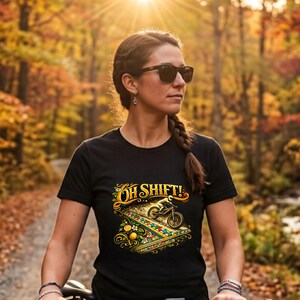 Könnte beinhalten: Schwarzes T-Shirt mit dem Text "Oh Shift!" und einer farbenfrohen Mountainbike-Grafik. Das Bild zeigt eine Frau mit Sonnenbrille, die ein Fahrrad mit einem Rennnummernschild hält. Ein weißer Helm hängt am Lenker.