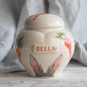 May include: A ceramic jar with a lid, featuring a speckled cream-colored finish. The jar is decorated with orange carrots and bunny ears. The name "BELLA" is displayed on a rectangular plaque. The jar is likely for pet treats or storage.