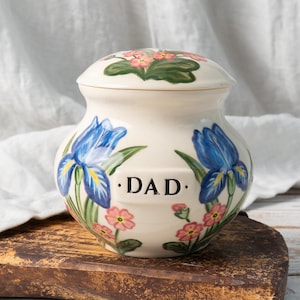 May include: A cream-colored ceramic jar with a lid, featuring hand-painted blue iris flowers and small pink blossoms. The word DAD is printed in black capital letters on the front. The jar has a rounded body and a matching domed lid.