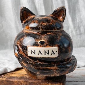 Può includere: Un barattolo in ceramica a forma di gatto con una finitura lucida maculata nera e marrone. Il gatto ha orecchie a punta e una coda arricciata che avvolge la parte anteriore. Una piccola placca rettangolare bianca al centro riporta NANA.