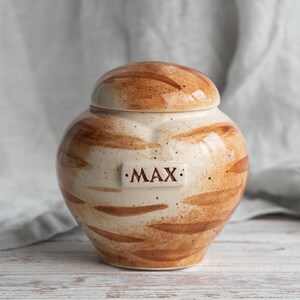 May include: A ceramic urn with a lid, cream-coloured with brown brushstroke patterns. The word "MAX" is displayed on a rectangular plaque. The urn is set against a neutral background.